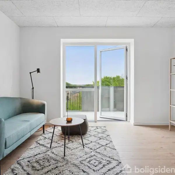 Sofa står i stuen med en åben altandør som baggrund. Stuen er lys med en reol til højre og et tæppe på gulvet.