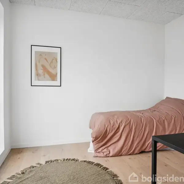 Seng med rosa sengetøj står mod en hvid væg med et kunsttryk. Rundt tæppe ligger på trægulv. Et bord og stol er nær vinduet med udsigt og blomster.