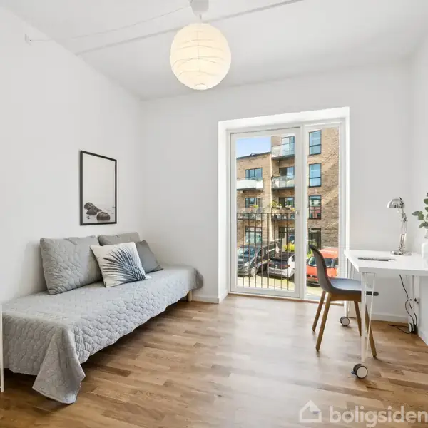 Sofa med gråt tæppe står ved en hvid væg med billede, lille bord ved siden. Skrivebord med stol står foran fransk altan. Gaden og bygninger kan ses udenfor.