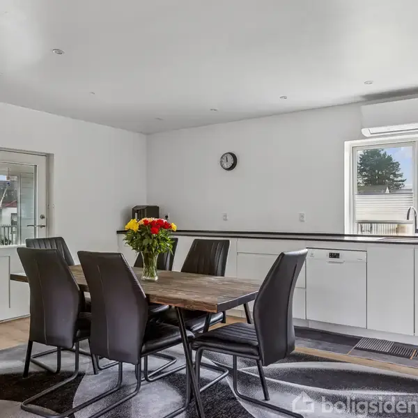 Et spisebord med seks sorte stole står i midten af et lyst køkken. Blomster i vase er på bordet. Store vinduer giver naturligt lys. En vægur hænger på væggen.