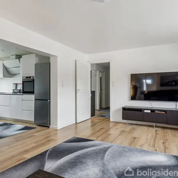 En stue med en fladskærm på en lav reol står stille. Rummet forbinder til et moderne køkken med spiseplads. Gulvet er lyst træ med sorte og grå tæpper.