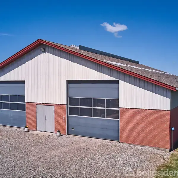Et stort lagerbygning med rødt tag og murstensvægge står på en grusbelagt grund, omgivet af græsplæne og træer under en blå himmel. Ingen synlig tekst.