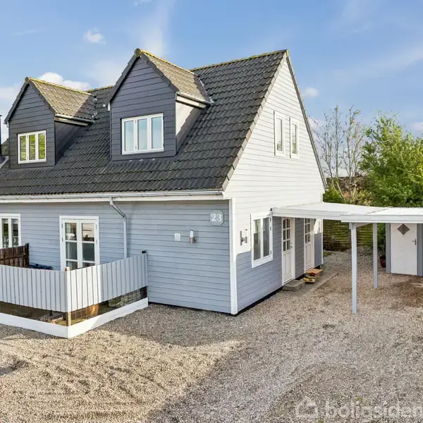 En gråmalet to-etagers hus med gavltag står i en grusbelagt gårdsplads. En carport ved siden af huset. Nummeret 23 vises på facaden.