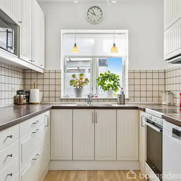 Køkkenet har hvide skabe, en mørk bordplade og hvide fliser. Et vindue med planter står åbent, mens en vægur hænger over. Gulvet er lyst træ, og der er diverse køkkenapparater.