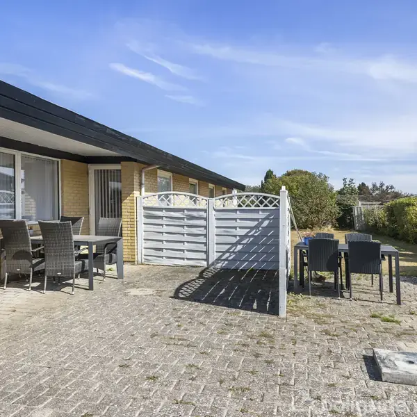Terrasse med havemøbler står på flisebelagt område. Solsiden af huset er murstensbeklædt. Skillevæg og grill til højre ses i nærhed af hæk. Klar himmel ovenfor.