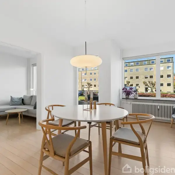 Et rundt spisebord med fire stole står i et lyst værelse. En lampe hænger over bordet. Store vinduer giver udsigt til en bygning og planter udenfor.