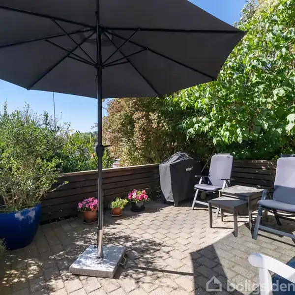 Parasollen skygger over terrasse med fliser, omgivet af træstakit. To stole og et bord står ved siden af farverige blomsterkrukker, med grønt løv som baggrund.