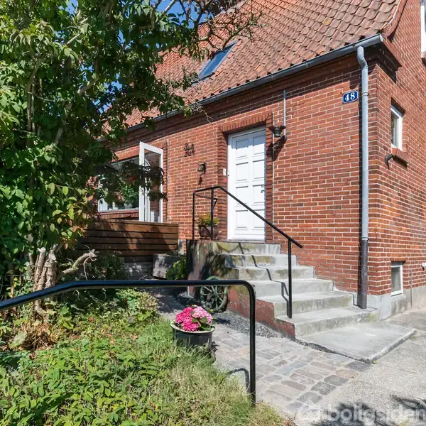 Rødt murstenshus med hvid dør og kvistvinduer. En trappe med sort gelænder leder op til døren. Omkringliggende grønne buske og blomster ved indgangen skaber en hyggelig atmosfære. Skilt: "143".