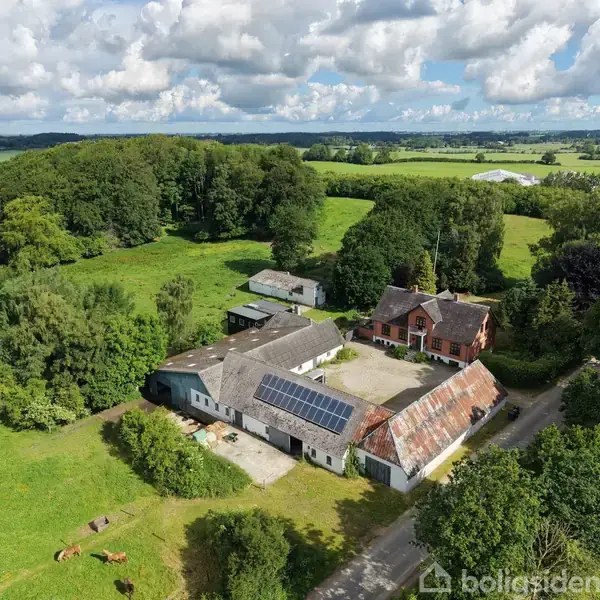 En landbrugsejendom med en hovedbygning og lagerhuse har solpaneler på taget. Den ligger omgivet af frodig grøn natur og marker under en let skyet himmel.