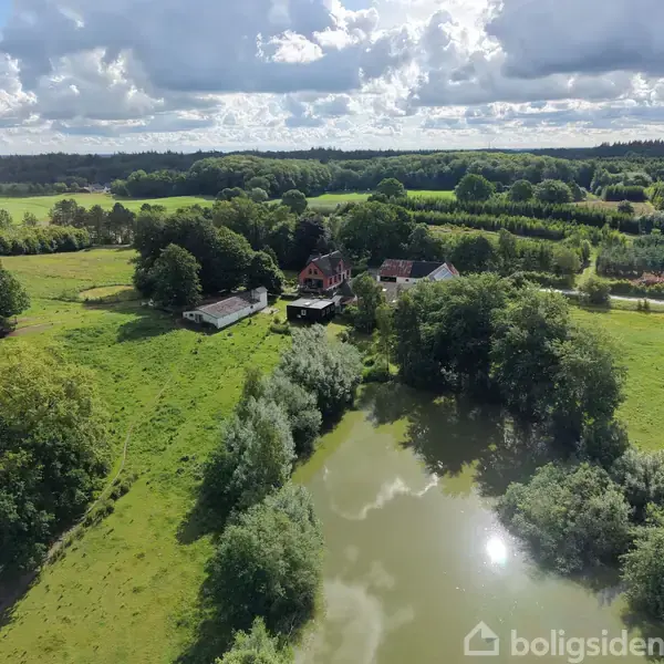 En gård med flere bygninger omgivet af træer og grønne marker. En sø ligger i forgrunden, og himlen er delvist skyet, hvilket skaber en fredelig landlig atmosfære.