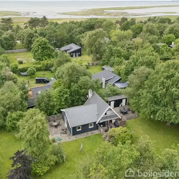 Et sort hus med gråt tag ligger omgivet af frodig, grøn vegetation. I baggrunden ses flere huse samt en kystlinje med vand.