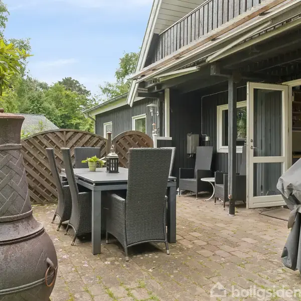 En udendørs terrasse med sorte havemøbler står ved siden af et sort hus med åbne døre. En stor grill og en haveovn er til stede. Omgivet af buske.