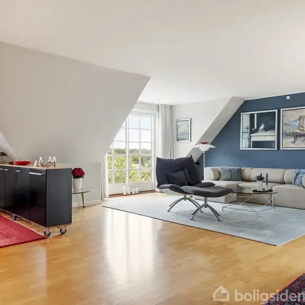 En lys stue har et hjørne med en beige sofa og et sofabord på et blåt tæppe. Et sort møbel står ved skråvæggen mod den åbne dør til en altan. Ingen tekst til stede.