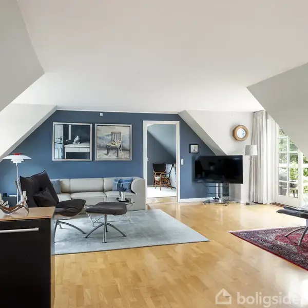 Stue har moderne møbler, herunder en sofa og lænestole på tæpper, med kunst på væggen og fladskærms-TV. Store vinduer giver udsigt til grøn natur udenfor.
