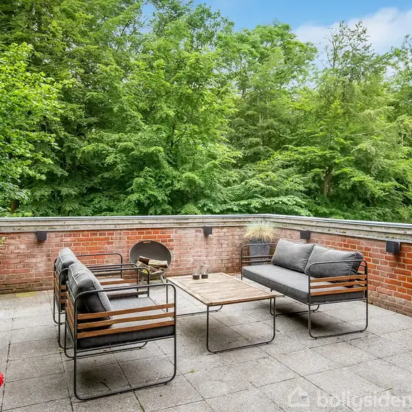 Terrassemøbler står stille på en flisebelagt terrasse med et lavt murværk. Omkring terrassen ses tætte, grønne træer, hvilket skaber en rolig, naturlig atmosfære.