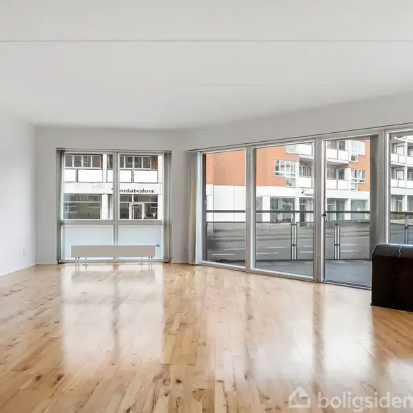 Et tomt værelse med trægulv har store vinduer og en glasdør ud til en altan. Udenfor ses tilstødende bygninger. En mørk træbænk eller kiste står til højre.