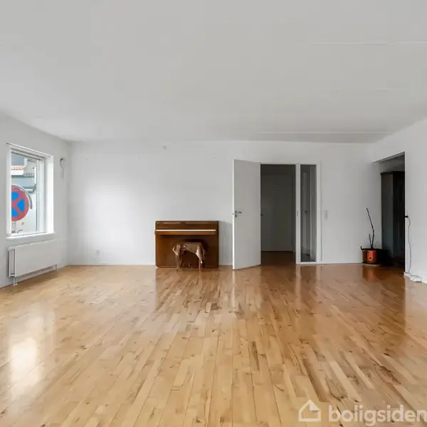 Et rummeligt, tomt rum med trægulv, et klaver ved væggen, og en åben dør fører til et tilstødende værelse. To store vinduer lader lys trænge ind.