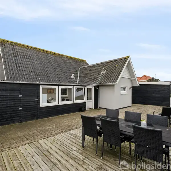 Hus med sort træfacade står stille på en træterrasse; omgivet af højt hegn med havemøbler i form af sort spisebord og otte stole foran huset.