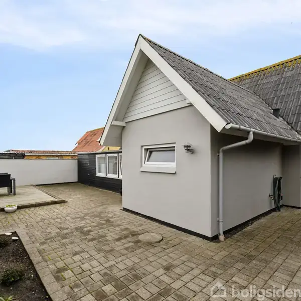Et moderne hus med spidse tage, malet i grå og hvid, står på en flisebelagt terrasse med havemøbler og beplantning i baggrunden under en klar, blå himmel.