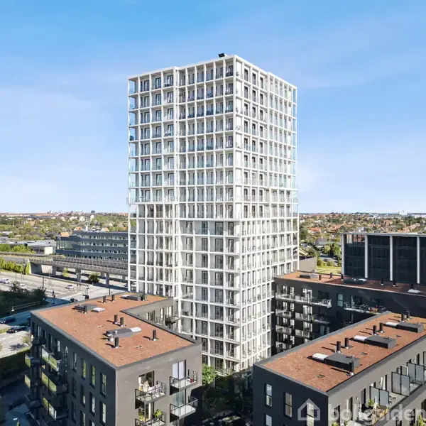 En høj, hvid moderne bygning med glasfacade står ved siden af lavere mørke bygninger. I baggrunden ses en motorvej samt en by med spredte grønne områder under en klar blå himmel.