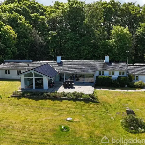 En hvid villa med sort tag står foran en frodig have. Villaen har store vinduer og terrasse. Bagved er en tæt skov med grønne træer.