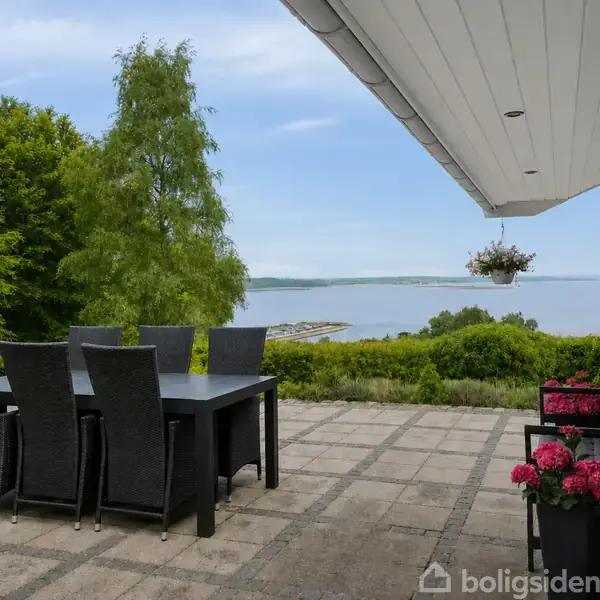 Et sort bord med seks stole står på en terrasse med fliser, tæt på et stort vindue. Smukke blomster er placeret i krukker, og der er udsigt til havet og træer.