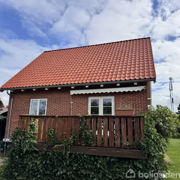 Hus med rødt tegltag og murstensvægge. En lille altan med trægelænder, omgivet af buske, befinder sig foran. Skyet himmel og grønt græs i forstadsområde.