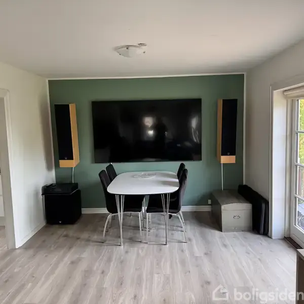 Fladskærms-tv hænger på en grøn væg, flankeret af højttalere. Under tv'et står et hvidt bord med sorte stole. Til højre ses en glasdør, der fører ud til en terrasse.