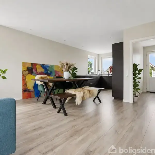 Træbord med fåreskindsovertrukne bænke står i moderne spisestue. Farverigt maleri læner sig mod væggen. Rummet er lyst med planter og store vinduer, der giver udsigt til himlen.
