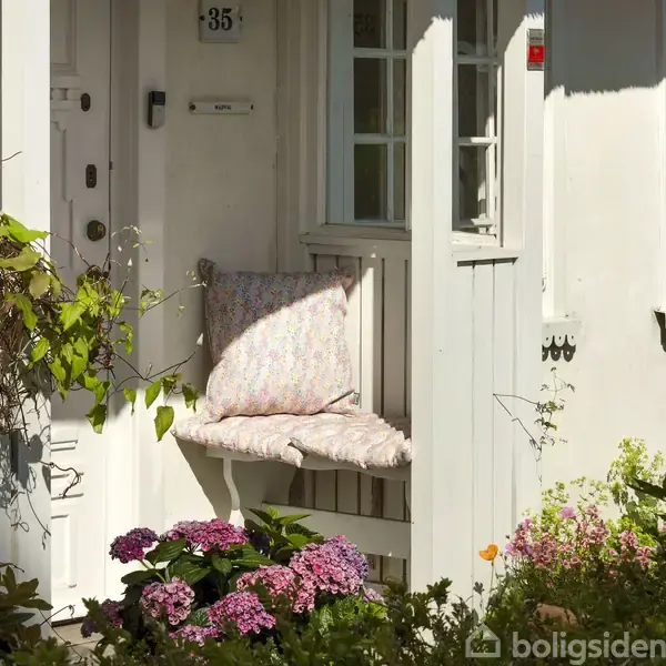 Hvidt hus med stråtag står i en velplejet have. Stensti snor sig gennem græsset mellem farverige blomster og buske. Hvidt hus ses i baggrunden.