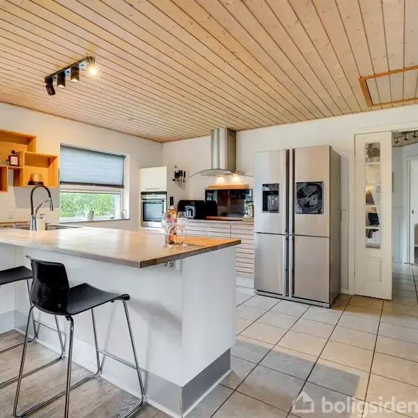 Køkken med lyst træloft, moderne apparater og en central køkkenø med to sorte barstole. Rummet har lyse fliser, åbne hylder og et stort vindue, der bringer naturligt lys ind.