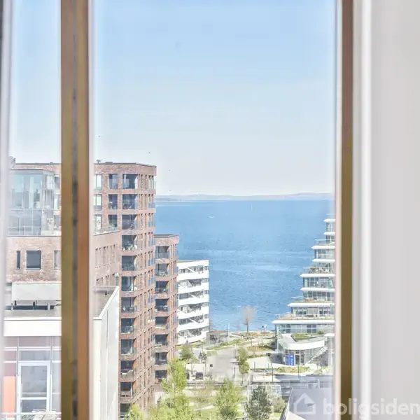 Udsigt gennem et vindue, viser havet i baggrunden omgivet af moderne lejlighedsbygninger i en bymæssig kystkontekst.