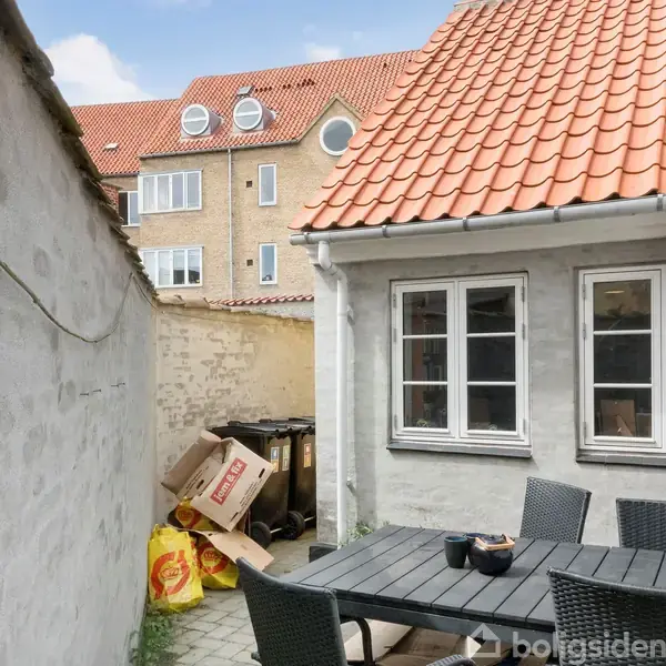 Terrassebord med sorte stole står i en lukket gårdhave ved et hus med rødt tegltag. Kartoner og poser er stablet op mod en mur. I baggrunden ses en bygning.