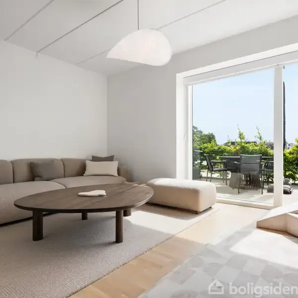 En beige sofa med puder står sammen med en lav, rund træbord i et lyst hjørne af en stue. To hvide vægge og et stort vindue rammer rummet ind.