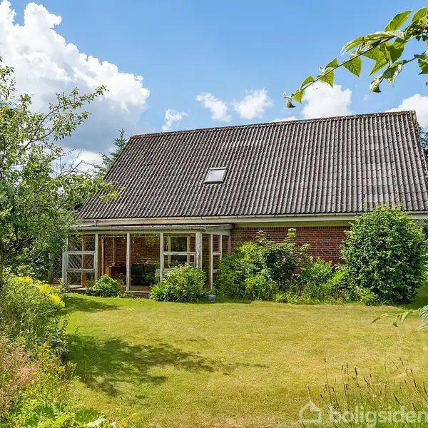 Hus med skråt tag står stille i en frodig have med grønt græs og tætte buske. Skyer ses på himlen i baggrunden.