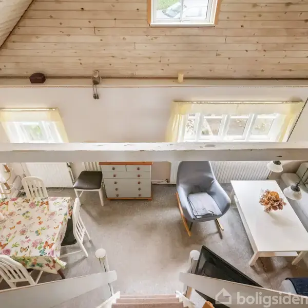 Loftbillede af en stue med spisebord dækket af en blomstret dug til venstre. Til højre findes en grå lænestol foran en sofa med sofabord, omgivet af lyse vægge og loft.