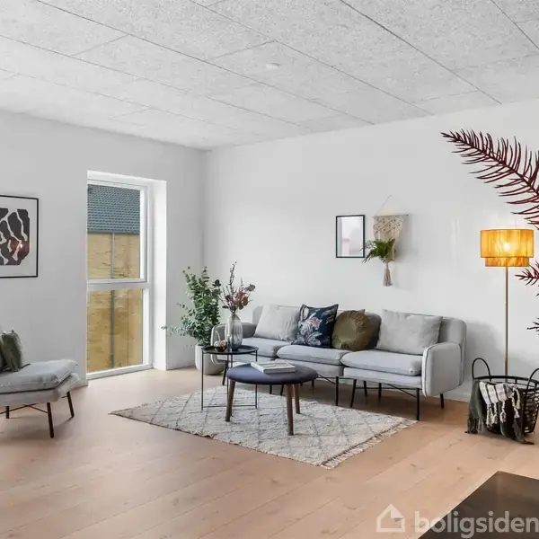 En moderne stue med grå sofa og lænestol på et lyst trægulv. Der er et rundt sofabord, planter, lamper og kunst på væggene. Store vinduer giver naturligt lys.