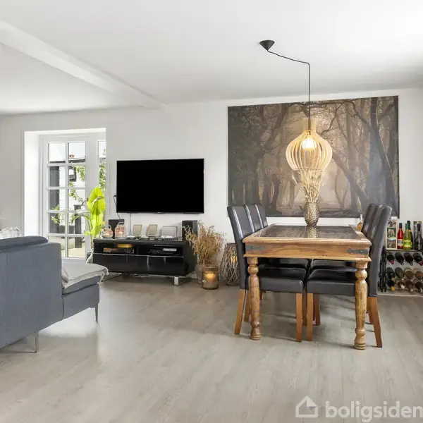 Stue med grå sofa, sofabord til venstre, og spisebord med lamper til højre. Fjernsyn placeret på reol. Stor vægdekoration bag spisebordet. Lysindfald fra vindue ved siden af planten.