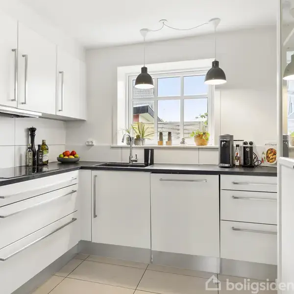 Et moderne hvidt køkken med sort bordplade og indbygget ovn. To lamper hænger over vasken ved et vindue med udsigt og planter. Bordet er dekoreret med frugter og køkkenredskaber.