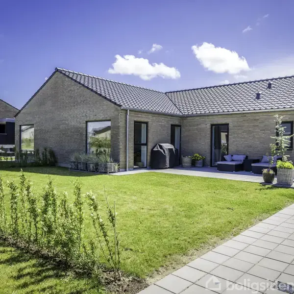Et moderne hus med stort glasparti og fladt tag står på en velplejet græsplæne. En terrasse med møbler er placeret ved siden af huset, under en klar blå himmel.