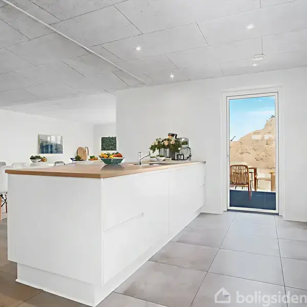 Køkken med hvidt køkkenbord og træoverflade, omgivet af spisestole og vindue ud mod terrasse. Indretning inkluderer planter og moderat belysning.