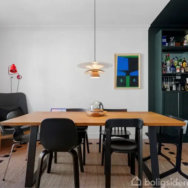 Et træbord fungerer som midtpunkt med seks stole omkring; en lampe hænger over bordet. Til højre er der en reol med flasker og glas. En åben dør til venstre.