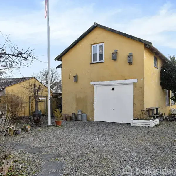 Et gult hus med garageport står på en grusbelagt gårdsplads, omgivet af en flagstang og buske. Huset har to små vinduer øverst og potteplanter foran.