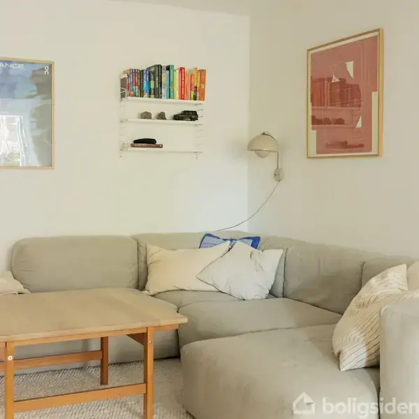 Grå hjørnesofa med puder står i en lys stue. Sofabord foran sofaen. Væghylde med bøger over sofaen. Kunstværker hænger på væggen sammen med en læselampe.