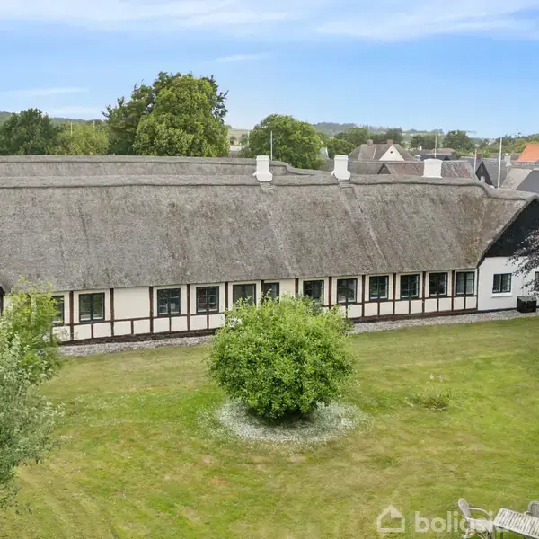 En lang stråtækt bygning står stille i midten af en grøn have med flere træer. Bagved er der udsigt til andre huse og en blå himmel.