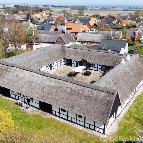 Bindingsværksgård med stråtag danner en gårdsplads med bord og parasol. Omkransende beboelseshuse og landskab med træer og græs. Marken omkranser den maleriske landsby.