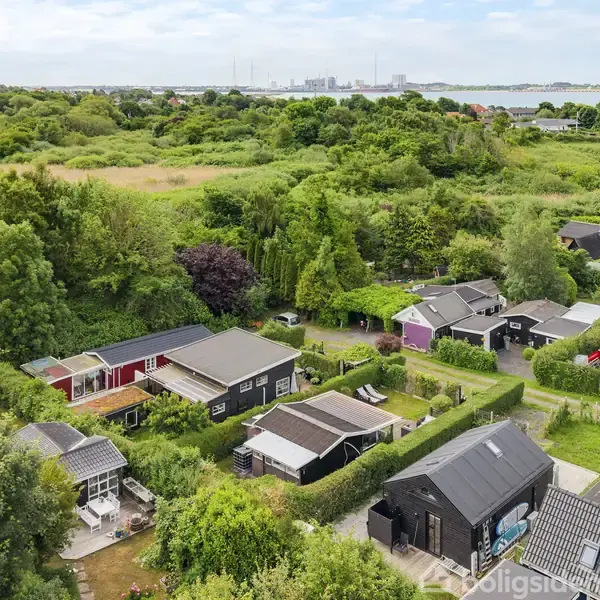 Huse i forskellige farver er placeret blandt frodige, grønne områder nær vandet. Landskabet strækker sig mod horisonten, hvor man kan ane skorstene og bygninger.