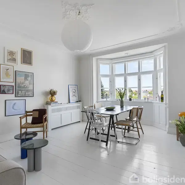 Stue med sofabord og sofa til venstre, spisebord med stole i midten nær stort vindue. Vægge prydet med diverse kunstværker. Lyse, minimalistiske omgivelser med planter og lyse farver.
