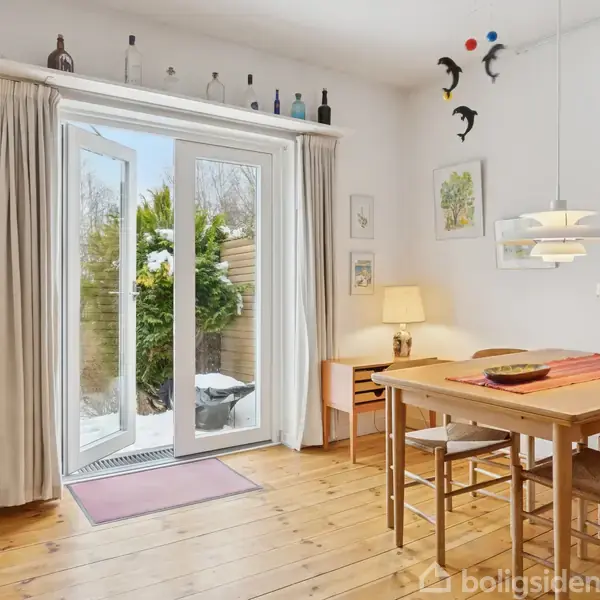 Spisestue med træbord og stole. Åbne glasdøre fører til en have. Vægkunst og lamper pynter rummet, mens flasker står på en hylde over dørene. En lille lampe står på et sidebord.