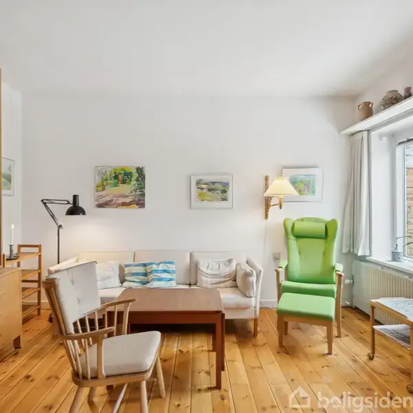 En stue med trægulv har en sofa, lænestol og flere stole omkring et sofabord. En bogreol fyldt med bøger står ved siden af. Væggene har kunstværker.